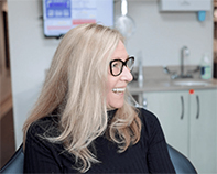 A woman with blonde hair, wearing glasses and a black top, is smiling at the camera while sitting in a dental chair.