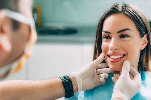 The image shows a woman seated in a dental chair with a smile, receiving dental care from a professional who appears to be a dentist, as indicated by the surgical mask and the presence of dental tools.