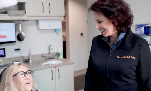 The image shows two individuals, a woman standing and smiling at the camera wearing a white coat with black text, and another woman seated in front of her, looking up towards the standing woman. They are both indoors, likely in a dental or medical office, as suggested by the equipment and setting behind them.