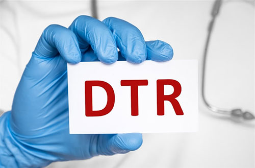 The image features a pair of gloved hands holding up a white card with the letters DTR in red, against a blurred background that appears to be a medical setting.