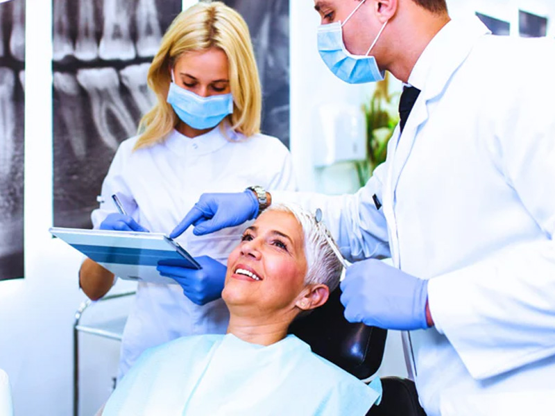 The image depicts a dental office setting where a woman is seated in the chair, receiving dental care from a professional wearing a face mask and using a device on her head.