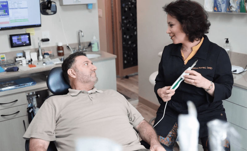 The image shows a dental office setting with two individuals one is seated in the dentist s chair while another stands by, possibly a dental professional.